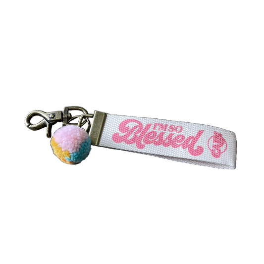 A keychain with a fabric strap that has the text 'Blessed' printed on it, along with a multicolored tassel.